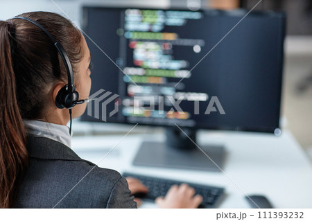 Over shoulder view of young woman wearing headset and using computer with code lines on screen in IT support office copy space 111393232