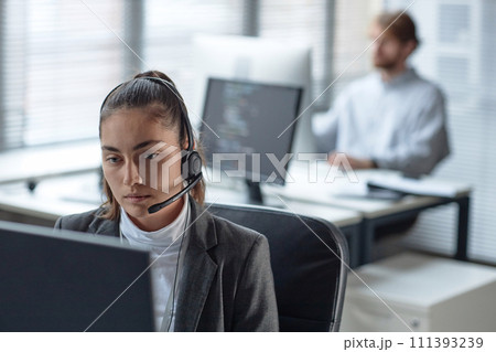Portrait of young woman wearing headset with microphone and using computer while working in customer support office copy space 111393239