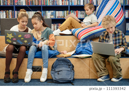 Full length view at group of young children enjoying break in modern library lounge in school and using computers Full length view at group of young children enjoying break in modern library lounge in school and using computers 111393342