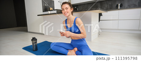 Sport and wellbeing. Young sportswoman does exercises at home, sits in yoga pose, holds smartphone, follows online training videos during her workout indoors Sport and wellbeing. Young sportswoman does exercises at home, sits in yoga pose, holds smartphone, follows online training videos during her workout indoors 111395798
