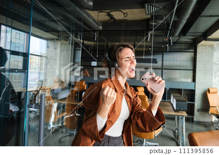 Portrait of businesswoman in office, singing, listening music in wireless headphones, having fun at workplace 111395856