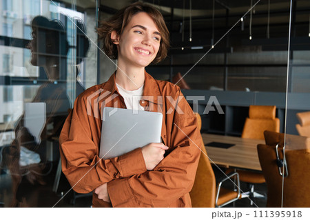 Portrait of young female manager, office worker with laptop, digital nomad programmer working at her workplace, standing with computer and smiling Portrait of young female manager, office worker with laptop, digital nomad programmer working at her workplace, standing with computer and smiling 111395918