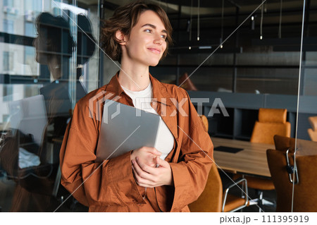 Portrait of young female manager, office worker with laptop, digital nomad programmer working at her workplace, standing with computer and smiling Portrait of young female manager, office worker with laptop, digital nomad programmer working at her workplace, standing with computer and smiling 111395919