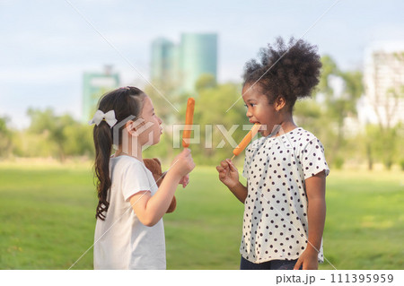 Happy cheerful white girls and black girl eat sausage together at outdoors park , Relationship little kids, Diverse ethnic concept. 111395959