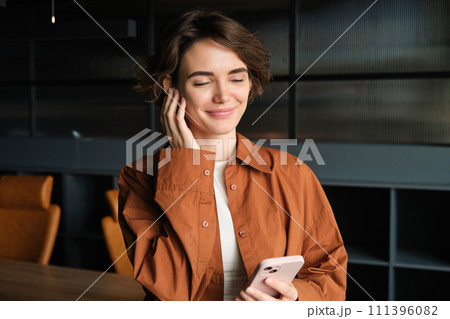 Young modern woman, office employee standing in conference room with smartphone and wireless headphones Young modern woman, office employee standing in conference room with smartphone and wireless headphones 111396082