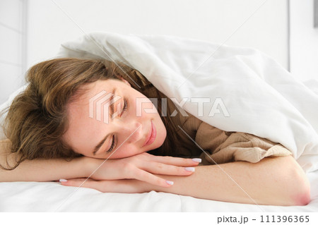 Portrait of happy young woman sleeping in her bed, enjoying a nap in hotel suite, lying under blanket 111396365