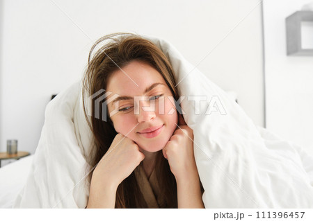 Cute girl with messy hair, lying in bed covered in white sheets duvet, smiling and laughing coquettish, spending time in her bedroom 111396457