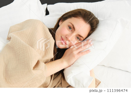 Portrait of gorgeous woman lying in bed, resting on pillow with eyes opened, looking at camera and smiling, wearing pyjamas, waking up in morning 111396474
