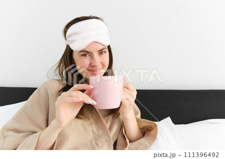 Happy young woman starting her morning with cup of coffee in bed, smiling and enjoying her lazy day, has sleeping mask on forehead Happy young woman starting her morning with cup of coffee in bed, smiling and enjoying her lazy day, has sleeping mask on forehead 111396492