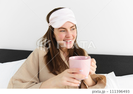 Happy young woman starting her morning with cup of coffee in bed, smiling and enjoying her lazy day, has sleeping mask on forehead 111396493