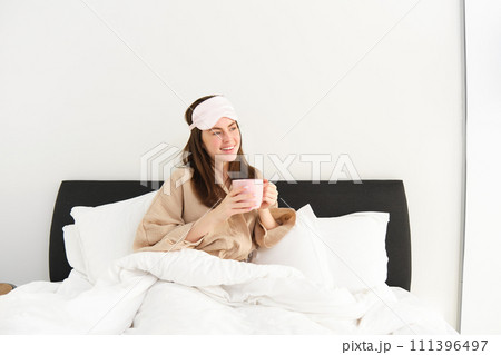 Portrait of beautiful woman in sleeping bag and pyjamas, drinking coffee in bed, enjoying her bright, relaxing morning 111396497