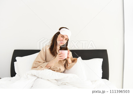 Portrait of beautiful woman relaxing in bed, waking up and having her morning cup of coffee in bedroom, looking happy 111396503