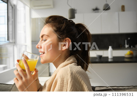 Close up portrait of happy young woman, enjoys relaxing day at home, drinks glass of orange juice and smiles, looks outside window 111396531