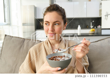 Relaxing morning at home. Young beautiful woman in bathrobe, eating cereals on sofa, having her breakfast in living room 111396553