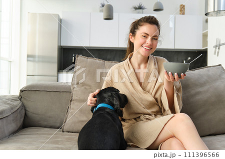 Happy mornings. Beautiful woman enjoying her morning, sitting on sofa, eating breakfast and playing, cuddling with dog, puppy asks for small bite of cereals Happy mornings. Beautiful woman enjoying her morning, sitting on sofa, eating breakfast and playing, cuddling with dog, puppy asks for small bite of cereals 111396566