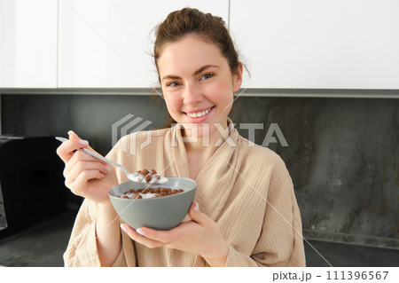 Gorgeous woman starts her day with cereals, eats breakfast and smiles, wears bathrobe, leans on kitchen worktop, starts her morning with quick meal Gorgeous woman starts her day with cereals, eats breakfast and smiles, wears bathrobe, leans on kitchen worktop, starts her morning with quick meal 111396567