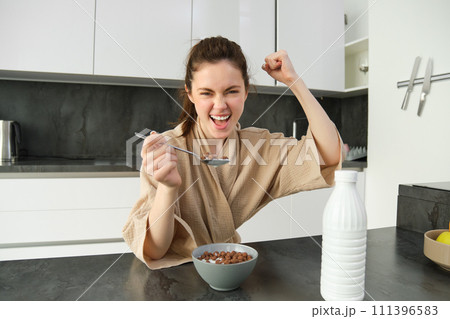 Portrait of enthusiastic young woman eating cereals with milk, looking excited and happy, sitting near kitchen worktop and having breakfast, raising hand up in triumph Portrait of enthusiastic young woman eating cereals with milk, looking excited and happy, sitting near kitchen worktop and having breakfast, raising hand up in triumph 111396583