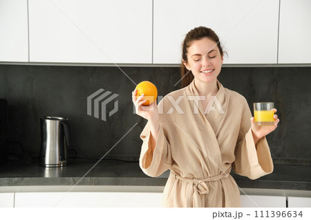 Image of good-looking healthy woman in bathrobe, drinking fresh juice, showing orange fruit, posing in kitchen Image of good-looking healthy woman in bathrobe, drinking fresh juice, showing orange fruit, posing in kitchen 111396634