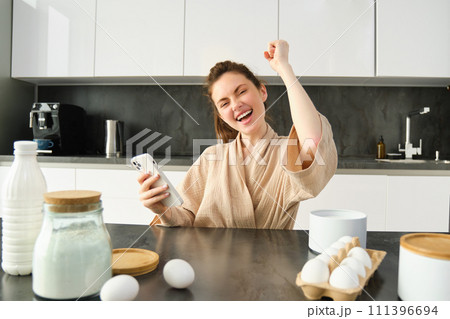 Attractive young cheerful girl baking at the kitchen, making dough, holding recipe book, having ideas 111396694