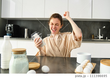 Attractive young cheerful girl baking at the kitchen, making dough, holding recipe book, having ideas Attractive young cheerful girl baking at the kitchen, making dough, holding recipe book, having ideas 111396695
