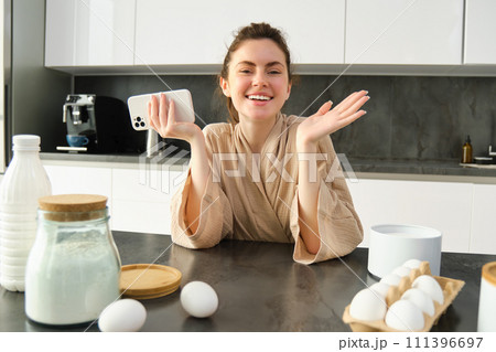 Attractive young cheerful girl baking at the kitchen, making dough, holding recipe book, having ideas 111396697