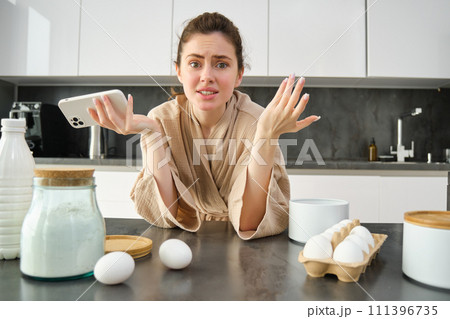Attractive young cheerful girl baking at the kitchen, making dough, holding recipe book, having ideas 111396735