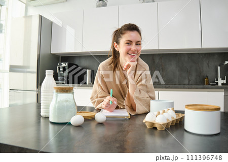 Attractive young cheerful girl baking at the kitchen, making dough, holding recipe book, having ideas Attractive young cheerful girl baking at the kitchen, making dough, holding recipe book, having ideas 111396748