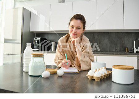 Attractive young cheerful girl baking at the kitchen, making dough, holding recipe book, having ideas Attractive young cheerful girl baking at the kitchen, making dough, holding recipe book, having ideas 111396753