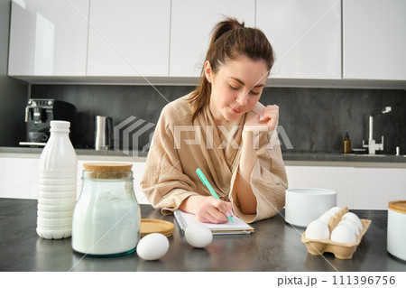 Attractive young cheerful girl baking at the kitchen, making dough, holding recipe book, having ideas 111396756