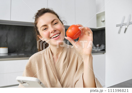 Close up portrait of beautiful smiling woman, holding fresh tomato, sitting in kitchen with smartphone, orders vegetables online, using application to buy groceries, using mobile phone 111396821