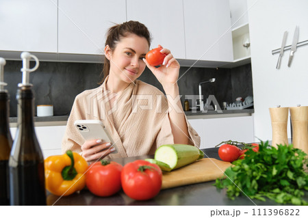 Image of young beautiful woman, holding tomato, sitting in kitchen with smartphone, chopping board and vegetables on counter, cooking food, order groceries for her recipe, using mobile phone app Image of young beautiful woman, holding tomato, sitting in kitchen with smartphone, chopping board and vegetables on counter, cooking food, order groceries for her recipe, using mobile phone app 111396822