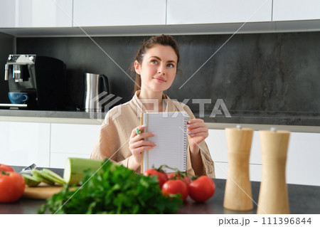 Portrait of happy young woman writes down menu for dinner, sits in the kitchen near vegetables, makes grocery list for shopping, poses in the bathrobe Portrait of happy young woman writes down menu for dinner, sits in the kitchen near vegetables, makes grocery list for shopping, poses in the bathrobe 111396844