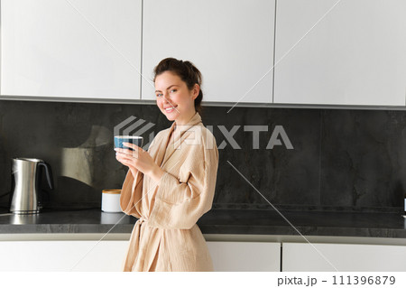 Daily routine and lifestyle. Young beautiful woman in bathrobe, standing in kitchen with cup of coffee, drinking tea, smiling and looking happy 111396879