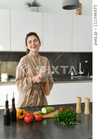 Vertical shot of young woman writing down food ideas, creating new meal, writing down grocery list or recipe in the notebook, standing in the kitchen near chopping board with fresh vegetables Vertical shot of young woman writing down food ideas, creating new meal, writing down grocery list or recipe in the notebook, standing in the kitchen near chopping board with fresh vegetables 111396975