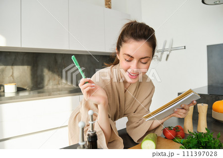 Portrait of woman checking grocery list, looking at vegetables, holding notebook, reading recipe while cooking meal in the kitchen, chopping tomatoes and zucchini 111397028