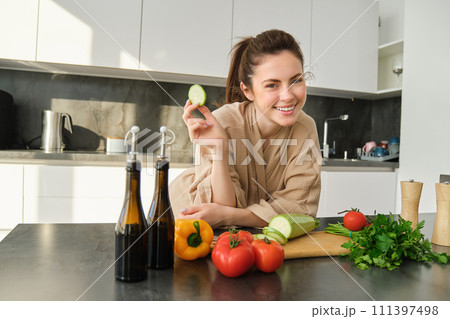 Portrait of happy girl eating vegetables while making meal, cooking healthy vegetarian food in kitchen, holding zucchini, posing in bathrobe Portrait of happy girl eating vegetables while making meal, cooking healthy vegetarian food in kitchen, holding zucchini, posing in bathrobe 111397498