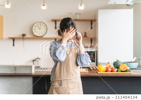 家のキッチンでスマホを持ちながら頭痛・体調不良・病気で泣く主婦のアジア人女性 家のキッチンでスマホを持ちながら頭痛・体調不良・病気で泣く主婦のアジア人女性 111404134