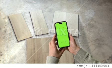 Hands Holding Device with Green Chroma Key Screen above Luxury Vinyl Wood Samples, Laminate On Floor In House. Choosing, Selecting Waterproof Flooring. Home Reconstruction. Horizontal Plane 111408903