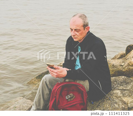 Midle aged man sitting on the shore of the lake. Solitude mature man using mobile phone on the bank. Concept of activ man 111411325