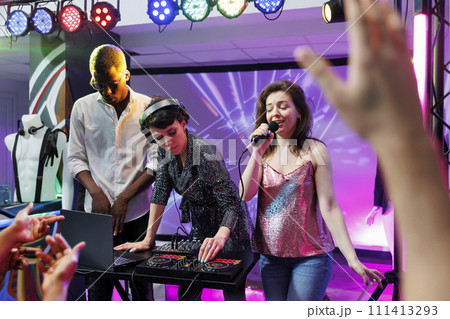 Dj and singer performing on stage at discotheque party in nightclub illuminated with colorful spotlights. Electronic music band singing and playing at disco gathering event in club 111413293