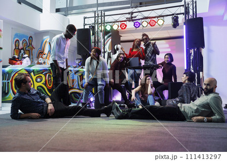 Music band performing on stage while diverse tired people lying and sitting on dancefloor. Exhausted young clubbers relaxing and listening to live music at club party gathering Music band performing on stage while diverse tired people lying and sitting on dancefloor. Exhausted young clubbers relaxing and listening to live music at club party gathering 111413297
