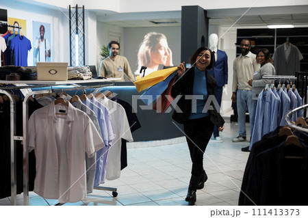 Clothing store satisfied customer walking with shopping bags in clothing store and looking at camera. Cheerful smiling asian woman carrying paper bags with trendy apparel purchase in mall Clothing store satisfied customer walking with shopping bags in clothing store and looking at camera. Cheerful smiling asian woman carrying paper bags with trendy apparel purchase in mall 111413373