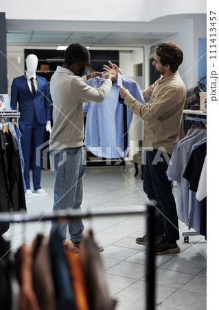 African american man selecting formal outfit and asking clothing store assistant for advice. Shopping mall boutique employee and customer discussing shirt and tie style match 111413457