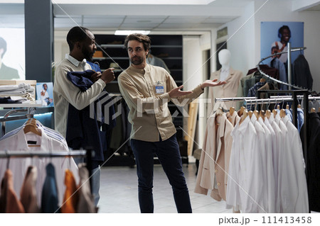 Shopping mall worker pointing at apparel rack while offering customer help in selecting casual outfit. Clothing store consultant assisting african american man in choosing garment 111413458