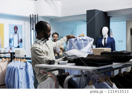 African american man checking shirt while exploring apparel rack in clothing store. Fashion boutique customer searching for formal garment while shopping in menswear section 111413513