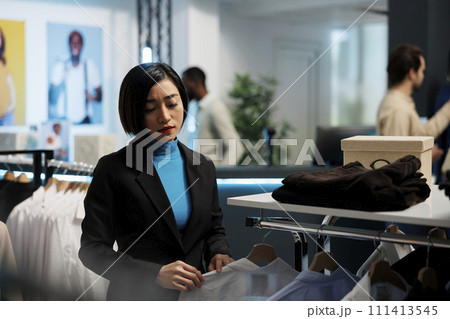 Young woman choosing fashionable clothes and browsing hanger in boutique. Stylish asian woman shopper examining garment on display rack while selecting outfit in trendy clothing store 111413545