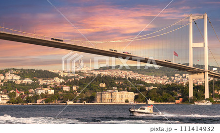 Sailboat traverses the Bosphorus Strait beneath the iconic Bosphorus Bridge, bathed in the warm glow of the setting sun 111414932