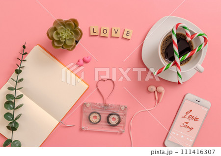 vintage transparent audio cassette magnetic tape in shape of heart, open blank book page, candy cane in the shape of heart on coffee cup, earphones, cactus, mobile phone playing love song on screen vintage transparent audio cassette magnetic tape in shape of heart, open blank book page, candy cane in the shape of heart on coffee cup, earphones, cactus, mobile phone playing love song on screen 111416307