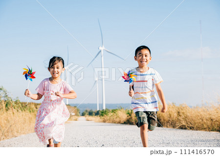 Brother and sister run by windmills holding pinwheels. Kids' joy near turbines represents a future of clean energy. Smiling children carefree and happy engage with sustainable technology. Brother and sister run by windmills holding pinwheels. Kids' joy near turbines represents a future of clean energy. Smiling children carefree and happy engage with sustainable technology. 111416570
