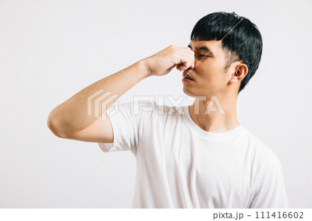 A disgusted Asian man holds his nose, expressing strong displeasure due to a bad smell situation. Studio shot isolated on white background, showcasing his humorous reaction. A disgusted Asian man holds his nose, expressing strong displeasure due to a bad smell situation. Studio shot isolated on white background, showcasing his humorous reaction. 111416602